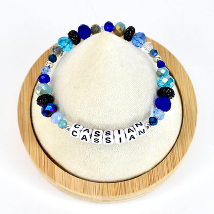 A beaded bracelet with the name "CASSIAN" spelled out in white letter beads. The bracelet features a variety of blue beads, including faceted, round, and crystal-like beads. The bracelet is displayed on a white surface.
