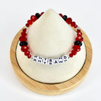 A beaded bracelet with red and black beads. The bracelet has the word "RHYSAND" spelled out in white letter beads. The bracelet is displayed on a white surface.