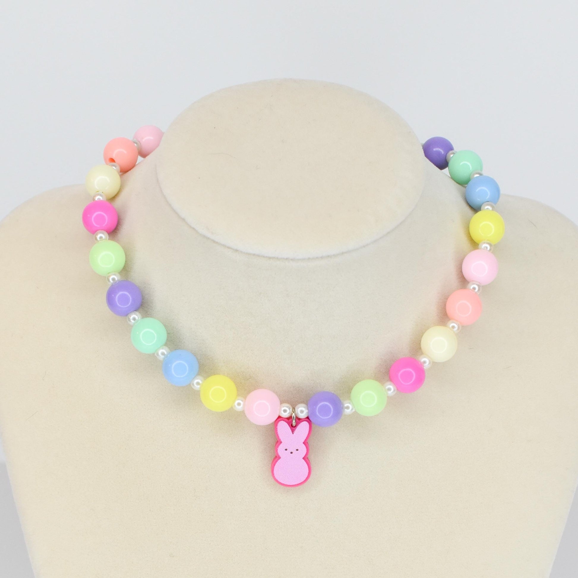 A colorful beaded necklace with pastel-colored beads in shades of pink, yellow, green, blue, and purple. The necklace features a pink bunny pendant. The necklace is displayed on a mannequin.