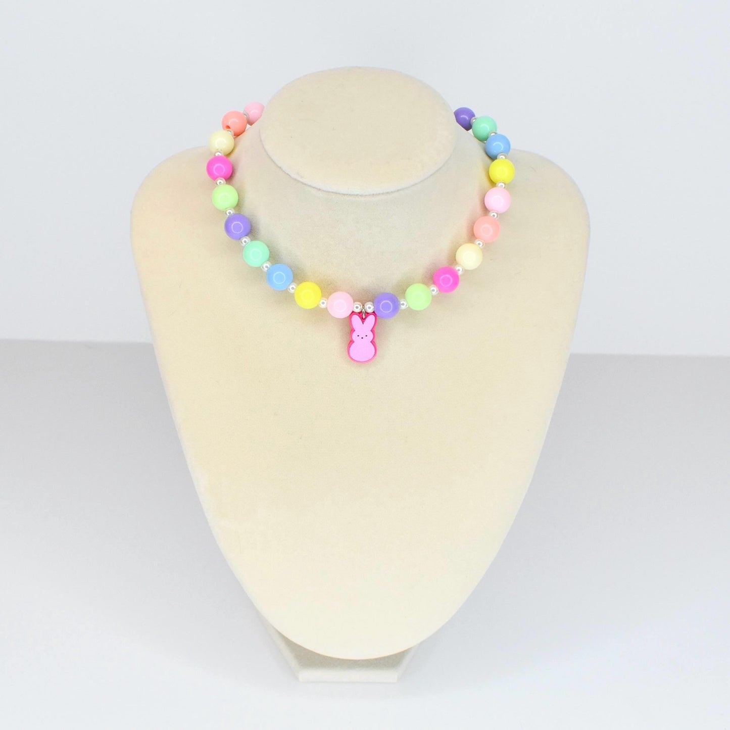 A colorful beaded necklace with pastel-colored beads in shades of pink, yellow, green, blue, and purple. A pink bunny charm hangs from the center. The necklace is displayed on a white mannequin.