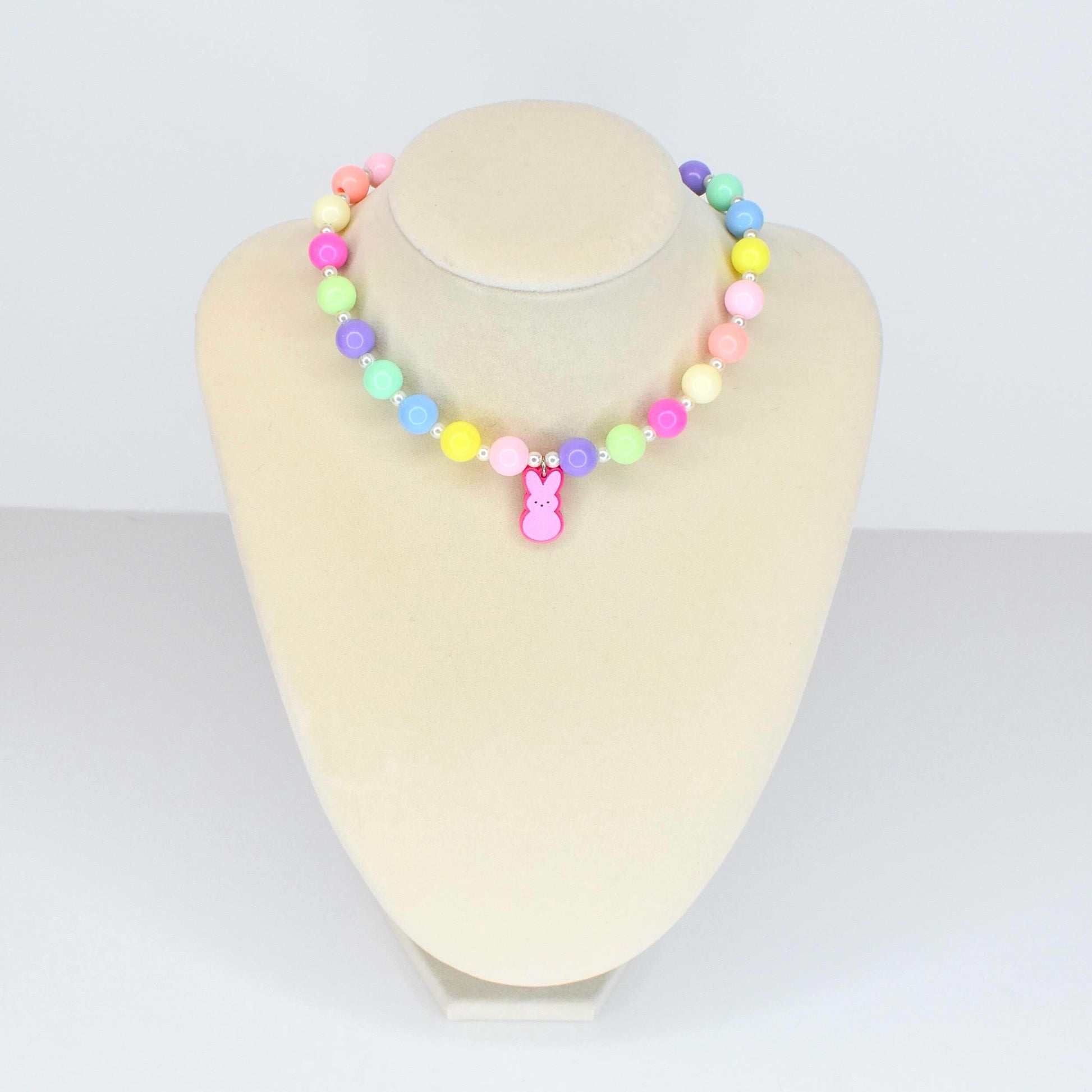 A colorful beaded necklace with pastel-colored beads in shades of pink, yellow, green, blue, and purple. A pink bunny charm hangs from the center. The necklace is displayed on a white mannequin.
