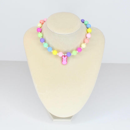 A colorful beaded necklace with pastel-colored beads in shades of pink, yellow, green, blue, and purple. A pink bunny charm hangs from the center. The necklace is displayed on a white mannequin.