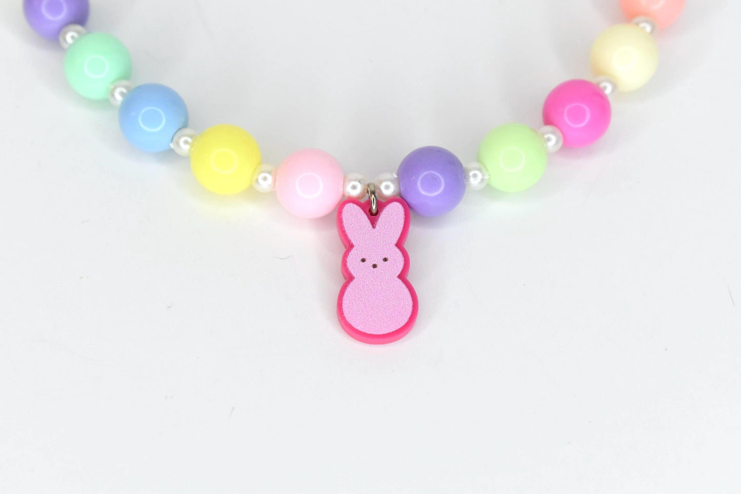 A colorful beaded necklace with a pink bunny charm. The necklace features large, round beads in pastel colors, including pink, yellow, blue, green, and purple, interspersed with small white beads. The bunny charm is pink with a darker pink outline.