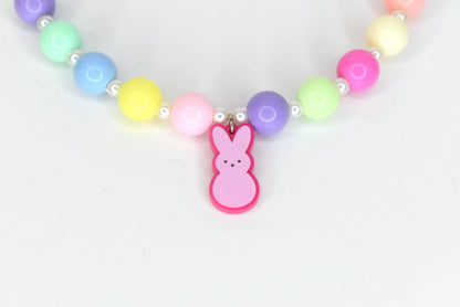 A colorful beaded necklace with a pink bunny charm. The necklace features large, round beads in pastel colors, including pink, yellow, blue, green, and purple, interspersed with small white beads. The bunny charm is pink with a darker pink outline.