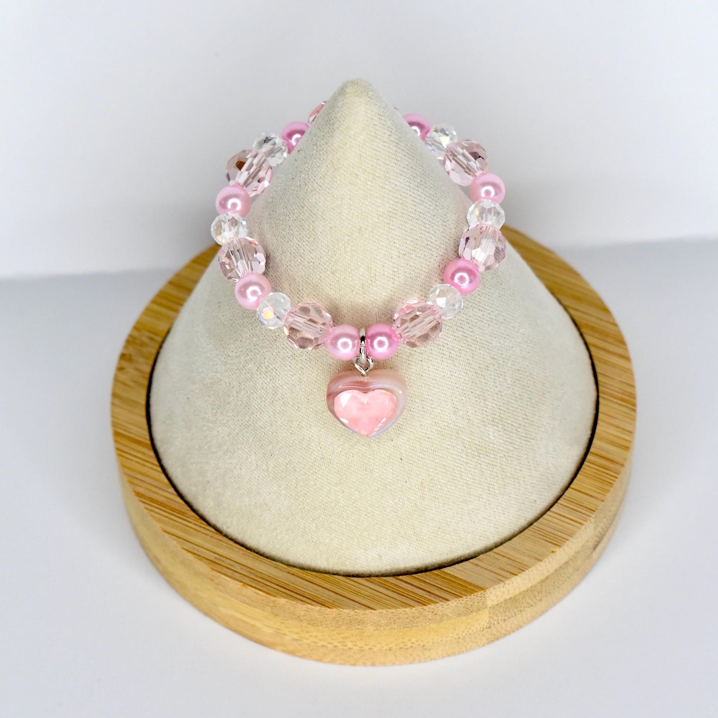 A pink beaded 17cm monster doll necklace with a heart charm. The necklace features pink and clear beads, with a small pink heart pendant. The necklace is displayed on a white surface and a wooden base.