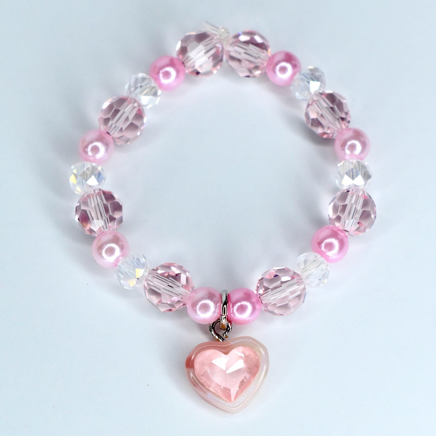 A pink beaded doll necklace with a heart charm. The necklace features a mix of pink, clear, and iridescent beads. The heart charm is a light pink color and is attached to the necklace with a silver-colored clasp.