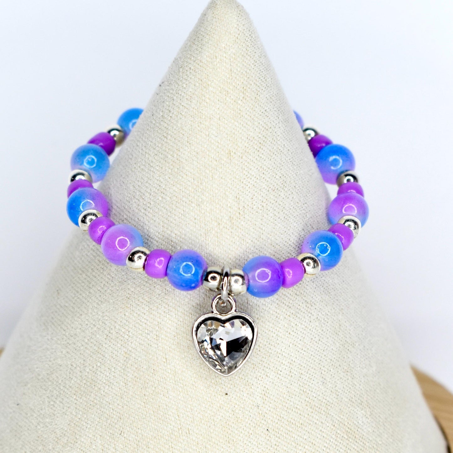 A beaded 17cm doll necklace featuring a heart-shaped charm. The necklace has blue, purple, and silver beads. The heart charm is clear and faceted. The necklace is displayed on a white surface.