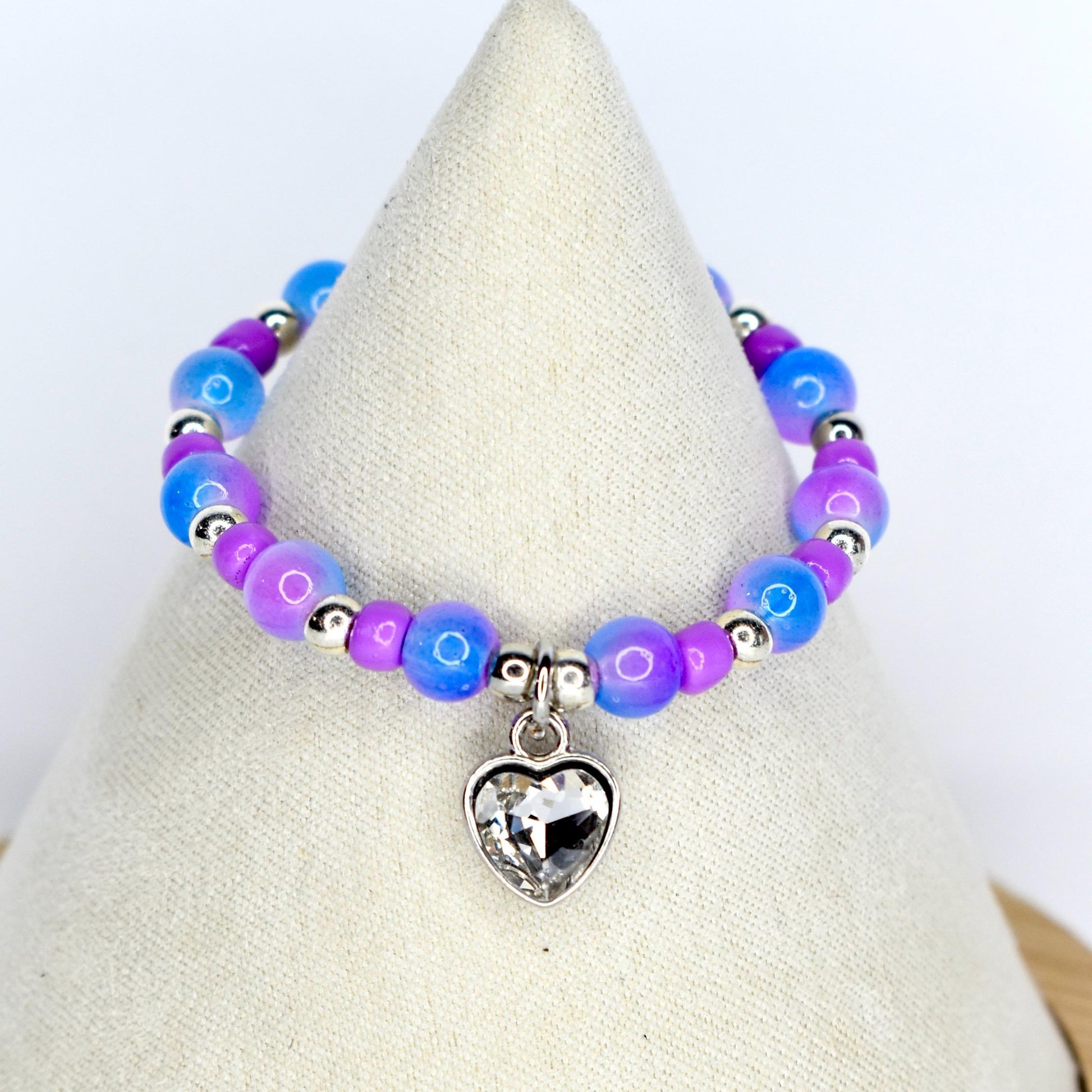 A beaded 17cm doll necklace featuring a heart-shaped charm. The necklace has blue, purple, and silver beads. The heart charm is clear and faceted. The necklace is displayed on a white surface.