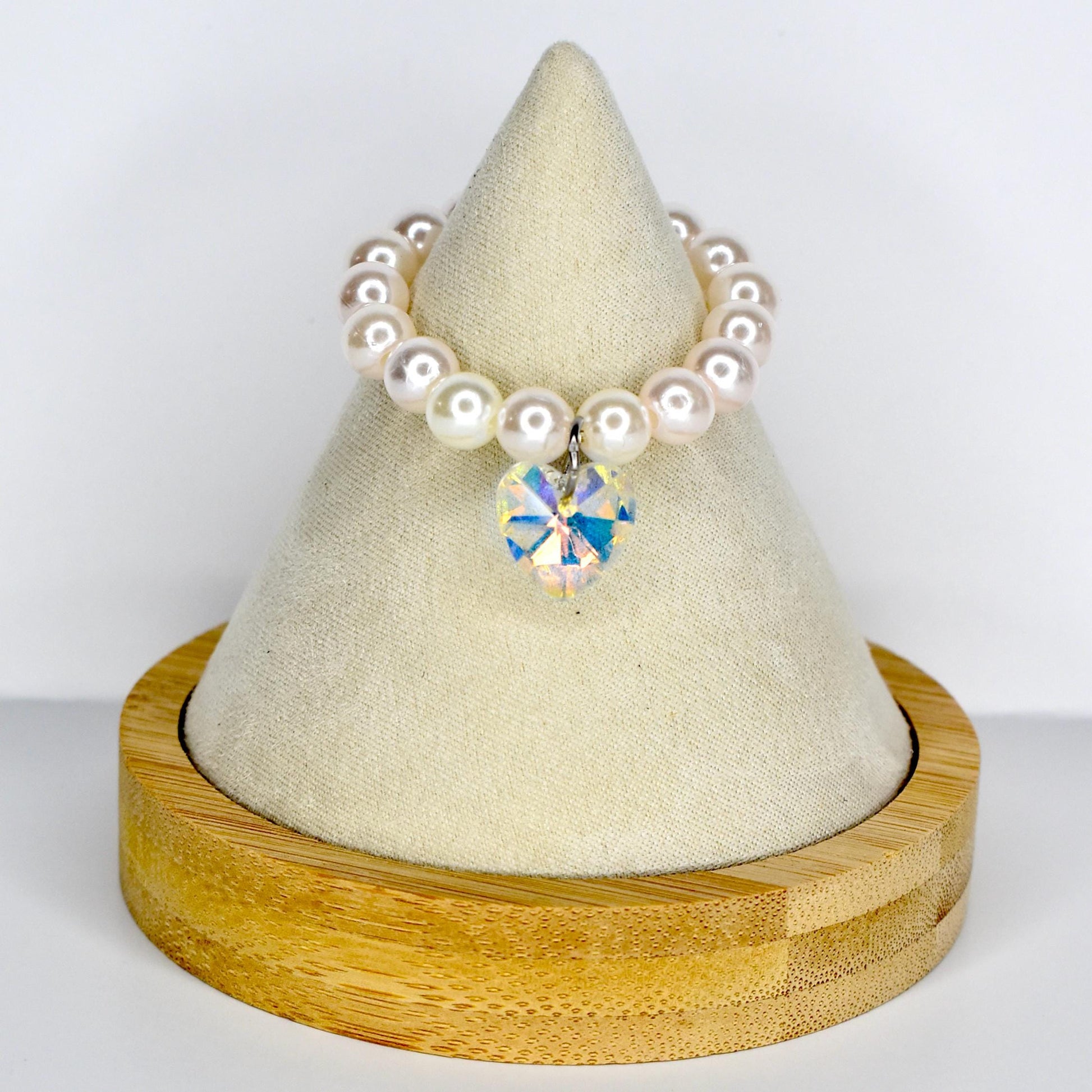 A pearl monster doll necklace with a heart-shaped iridescent crystal pendant. The necklace is made of round, white pearls. The pendant is a multi-faceted crystal that reflects various colors. The necklace is displayed on a white, cone-shaped stand with a wooden base.