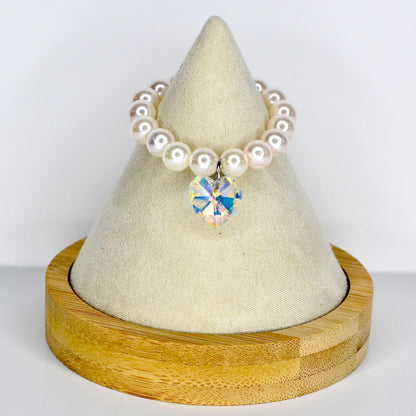 A pearl monster doll necklace with a heart-shaped iridescent crystal pendant. The necklace is made of round, white pearls. The pendant is a multi-faceted crystal that reflects various colors. The necklace is displayed on a white, cone-shaped stand with a wooden base.