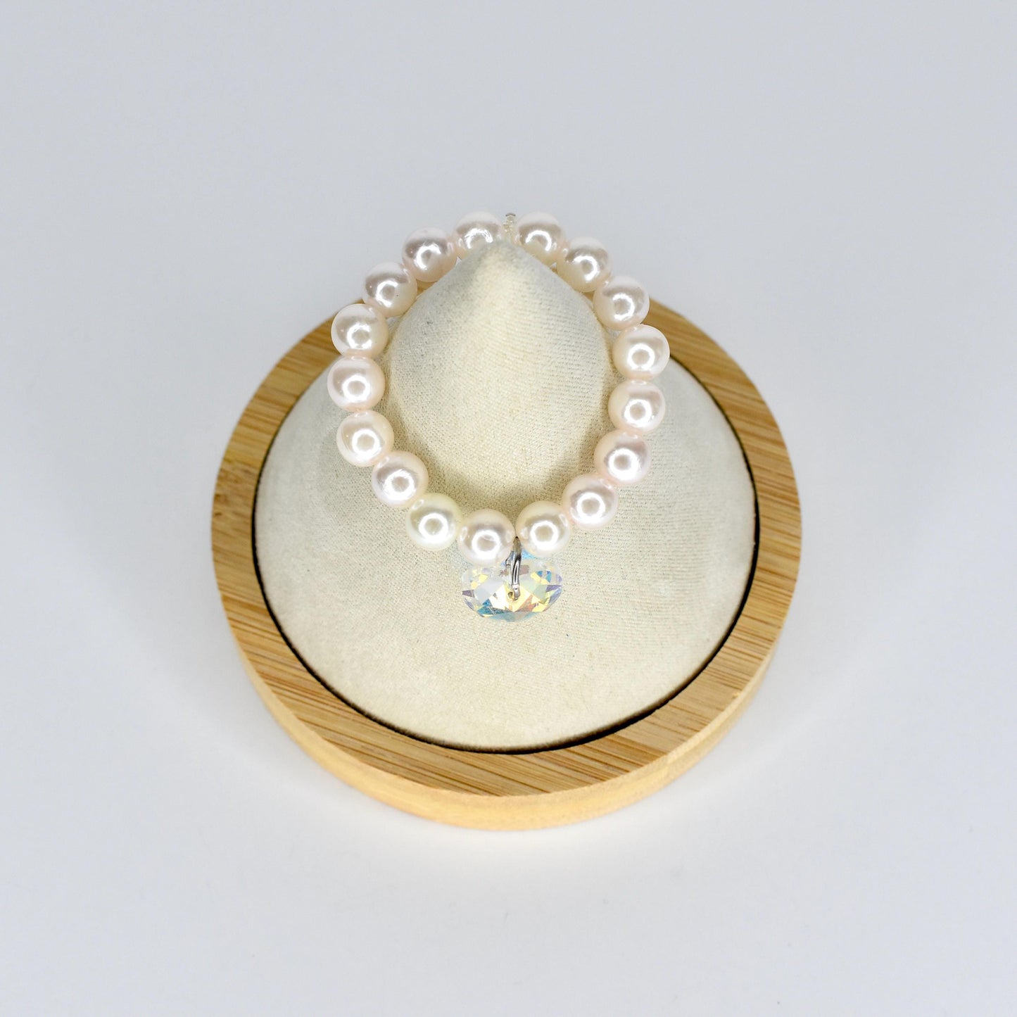 A pearl mini necklace with a clear crystal pendant. The necklace is made of round, white pearls. The crystal pendant is heart-shaped and reflects multiple colors. The necklace is displayed on a cream-colored cushion inside a wooden frame.