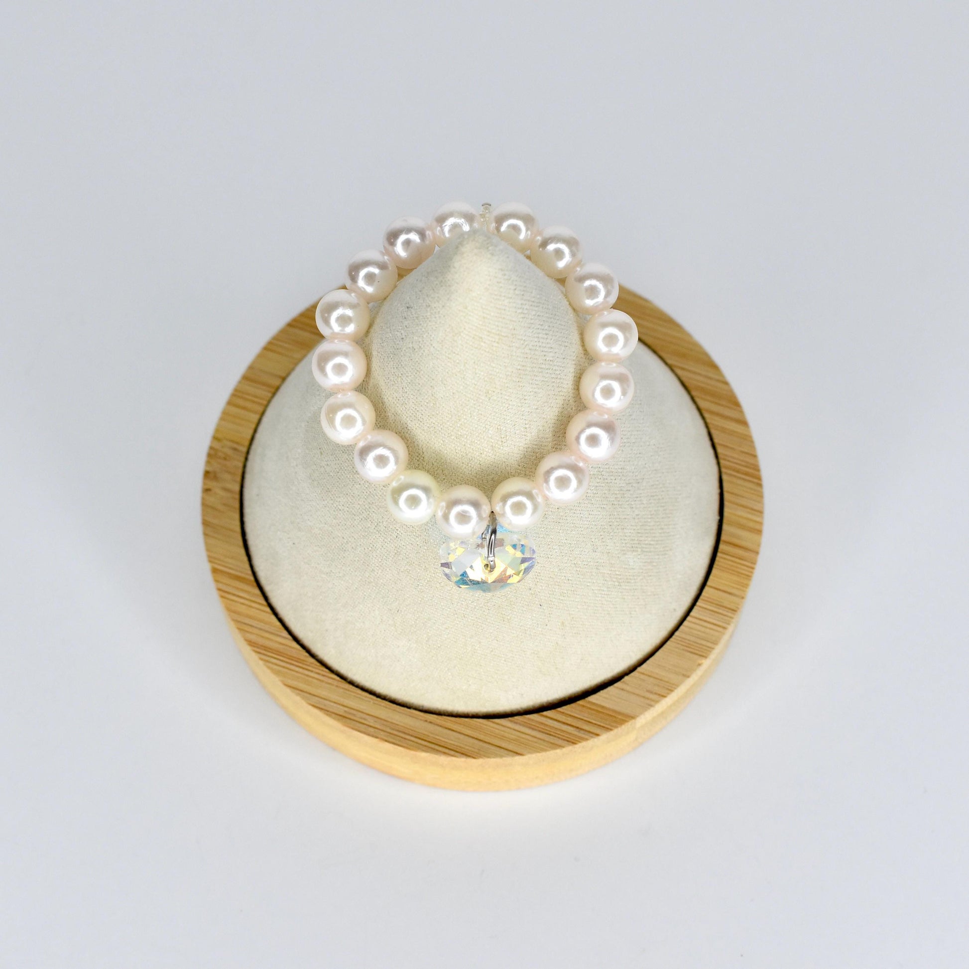 A pearl mini necklace with a clear crystal pendant. The necklace is made of round, white pearls. The crystal pendant is heart-shaped and reflects multiple colors. The necklace is displayed on a cream-colored cushion inside a wooden frame.