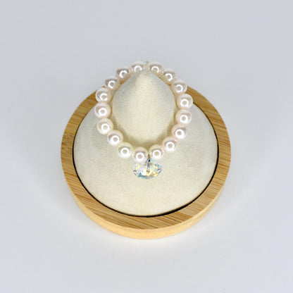 A pearl mini necklace with a clear crystal pendant. The necklace is made of round, white pearls. The crystal pendant is heart-shaped and reflects multiple colors. The necklace is displayed on a cream-colored cushion inside a wooden frame.