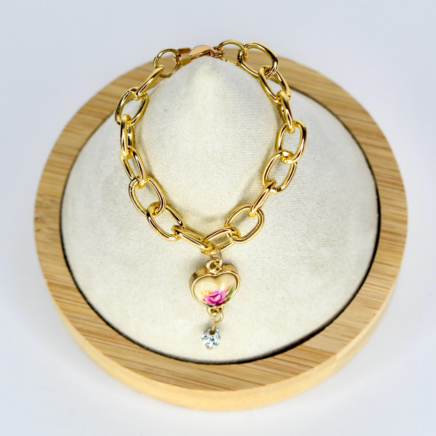 Gold-toned chain doll necklace with a heart-shaped charm. The charm features a floral design with pink and white roses. A small, clear crystal dangles below the heart. The necklace is displayed on a white surface within a wooden frame.