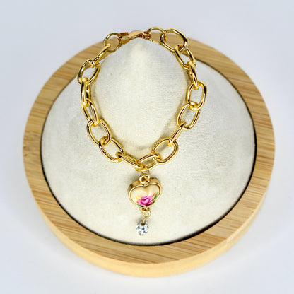 Gold-toned chain doll necklace with a heart-shaped charm. The charm features a floral design with pink and white roses. A small, clear crystal dangles below the heart. The necklace is displayed on a white surface within a wooden frame.