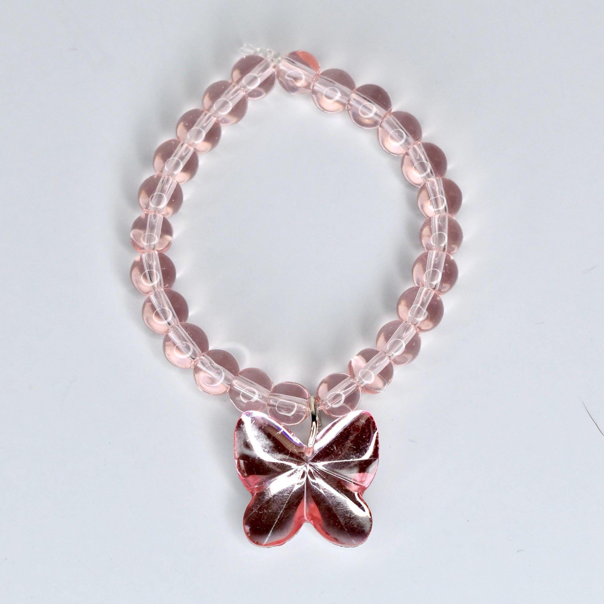 A pink beaded 17cm monster doll necklace with a butterfly pendant. The necklace features translucent pink beads strung together. The butterfly pendant is a darker shade of pink and has a reflective, faceted design. The necklace is designed to be worn around the wrist.