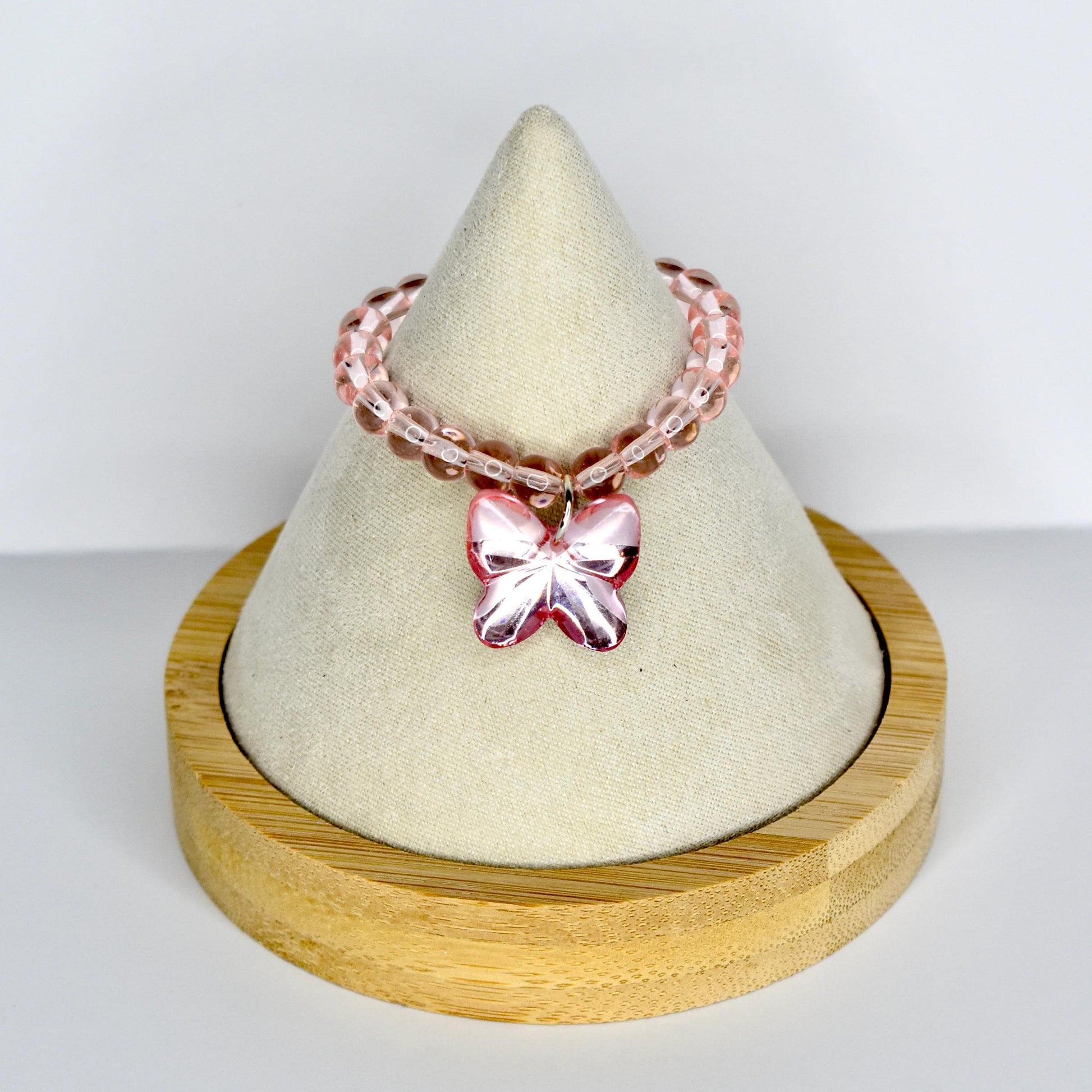 A pink beaded doll necklace with a butterfly charm. The necklace is displayed on a white cone-shaped stand, set on a round wooden base. The butterfly charm is a darker shade of pink.