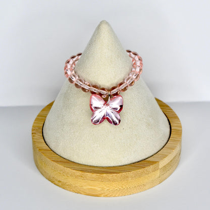 A pink beaded doll necklace with a butterfly charm. The necklace is displayed on a white cone-shaped stand, set on a round wooden base. The butterfly charm is a darker shade of pink.