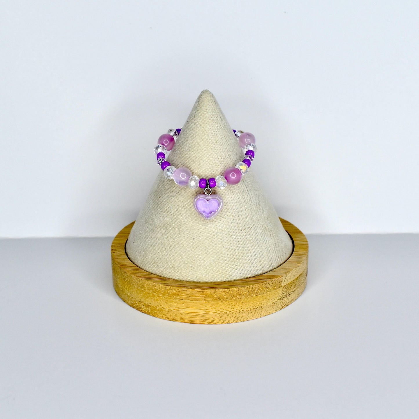 A beaded doll necklace featuring a variety of purple and clear beads. A small, light purple heart charm hangs from the center. The necklace is displayed on a cream-colored cone-shaped stand with a wooden base.