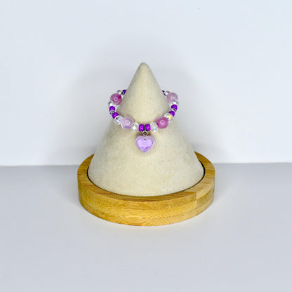 A beaded doll necklace featuring a variety of purple and clear beads. A small, light purple heart charm hangs from the center. The necklace is displayed on a cream-colored cone-shaped stand with a wooden base.