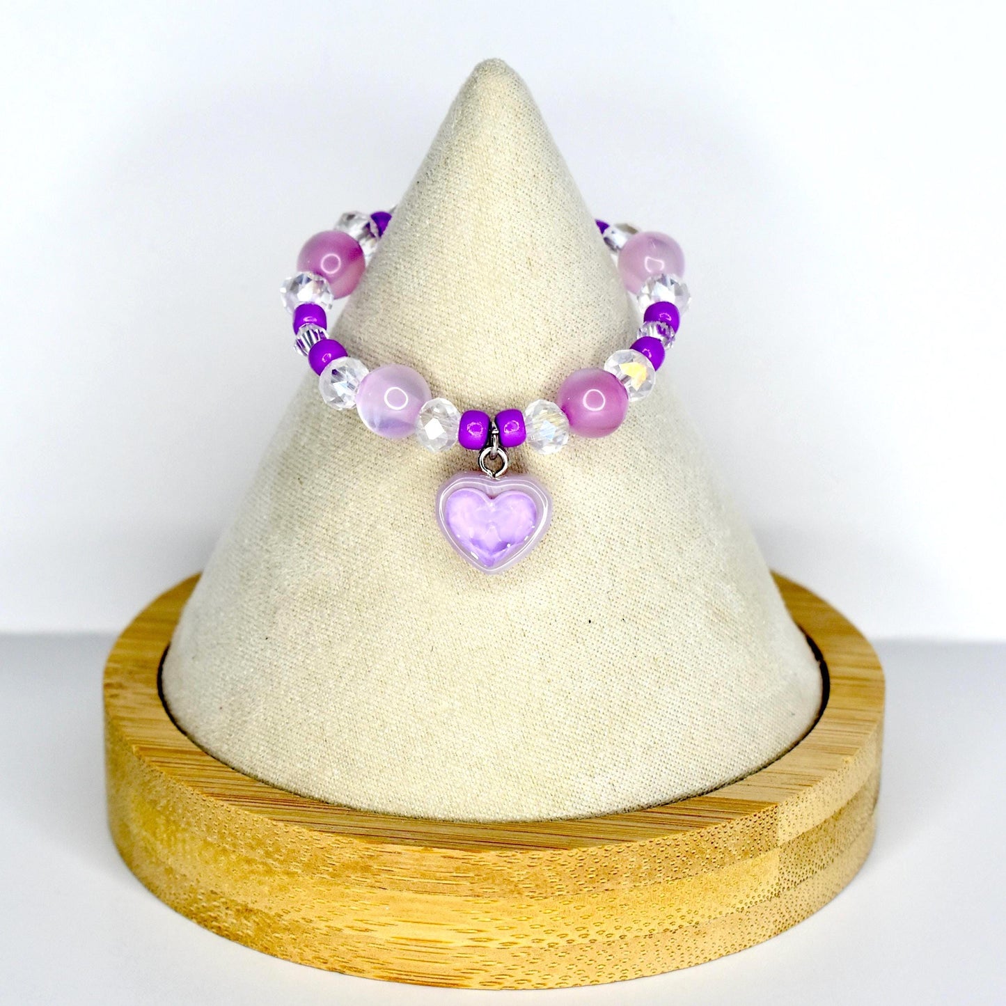 A beaded monster doll necklace featuring a lavender purple heart charm. The necklace has a mix of purple, clear, and iridescent beads. The heart charm is a light purple color. The necklace is displayed on a white cone-shaped stand with a wooden base.