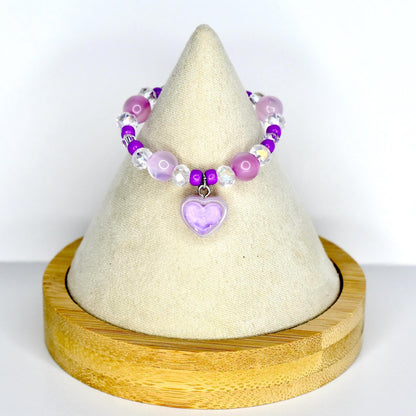 A beaded monster doll necklace featuring a lavender purple heart charm. The necklace has a mix of purple, clear, and iridescent beads. The heart charm is a light purple color. The necklace is displayed on a white cone-shaped stand with a wooden base.
