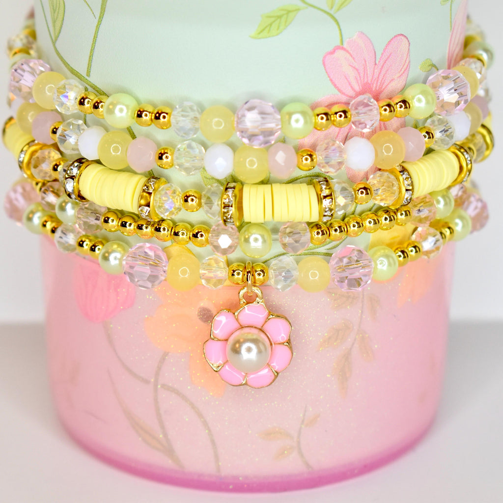 A stack of beaded bracelets in pastel colors. The bracelets feature yellow, pink, and clear beads, with gold accents. A pink flower charm with a pearl center hangs from the bottom bracelet. The bracelets are stacked on a pink and green floral-patterned container.