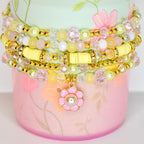 A stack of beaded bracelets in pastel colors. The bracelets feature yellow, pink, and clear beads, with gold accents. A pink flower charm with a pearl center hangs from the bottom bracelet. The bracelets are stacked on a pink and green floral-patterned container.