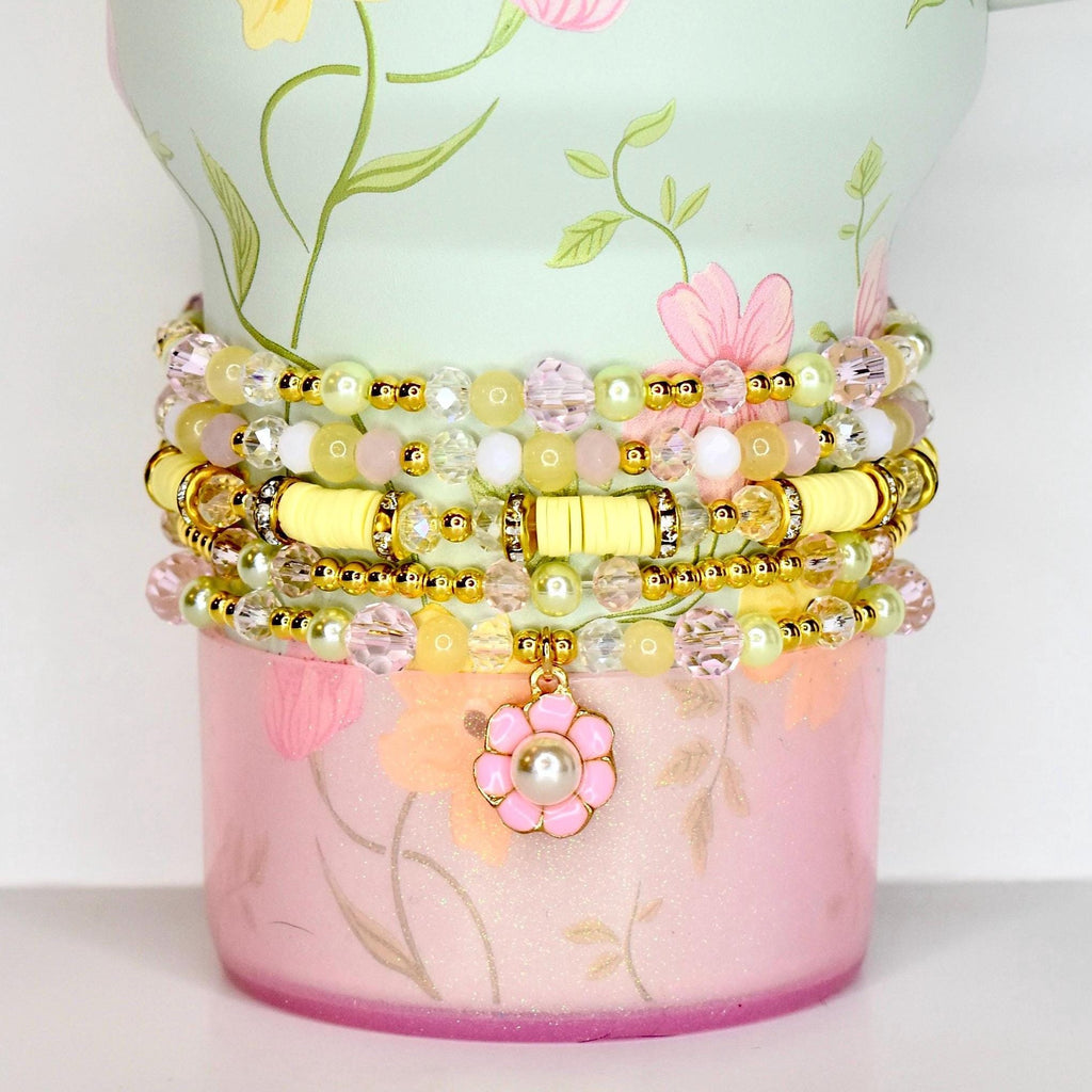 Stack of beaded bracelets in shades of yellow, pink, and clear. The bracelets are adorned with gold accents and a flower charm. The bracelets are displayed on a floral-patterned container.