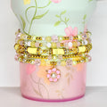 Stack of beaded bracelets in shades of yellow, pink, and clear. The bracelets are adorned with gold accents and a flower charm. The bracelets are displayed on a floral-patterned container.