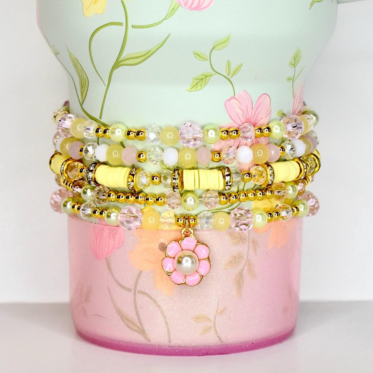 Stack of beaded bracelets in shades of yellow, pink, and clear. The bracelets are adorned with gold accents and a flower charm. The bracelets are displayed on a floral-patterned container.