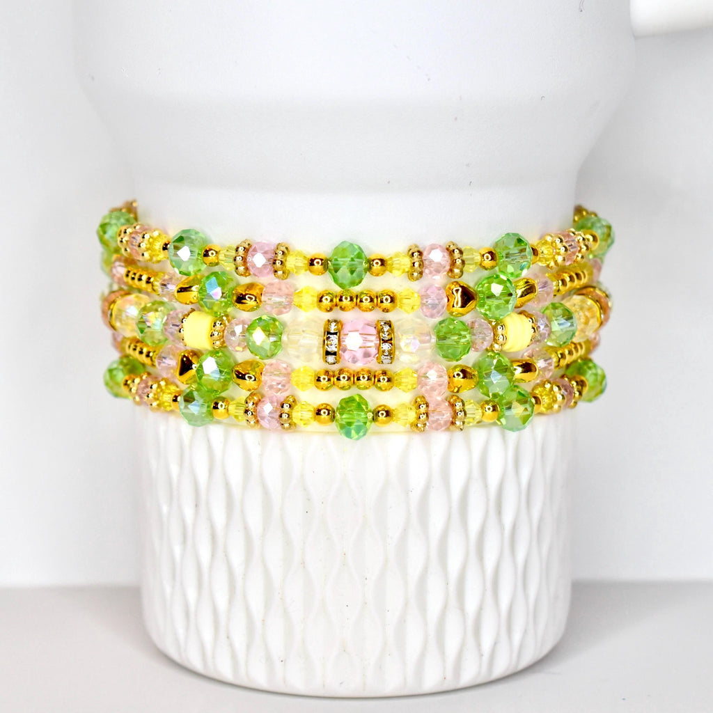 A stack of beaded bracelets in shades of yellow, pink, and green. The bracelets are adorned with gold-colored beads and clear, faceted beads. The bracelets are displayed on a white, textured surface.