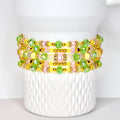 A stack of beaded bracelets in shades of yellow, pink, and green. The bracelets are adorned with gold-colored beads and clear, faceted beads. The bracelets are displayed on a white, textured surface.