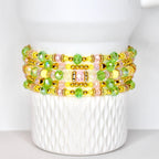 A stack of beaded bracelets in shades of yellow, pink, and green. The bracelets are adorned with gold-colored beads and clear, faceted beads. The bracelets are displayed on a white, textured surface.