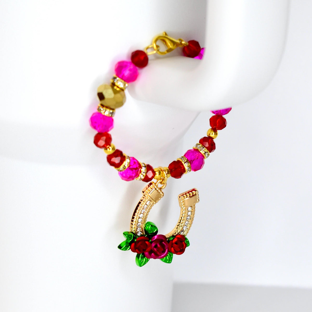 A beaded tumbler handle bracelet with a gold horseshoe charm. The bracelet features pink, red, and gold beads. The horseshoe charm is gold with clear crystals and red roses with green leaves.