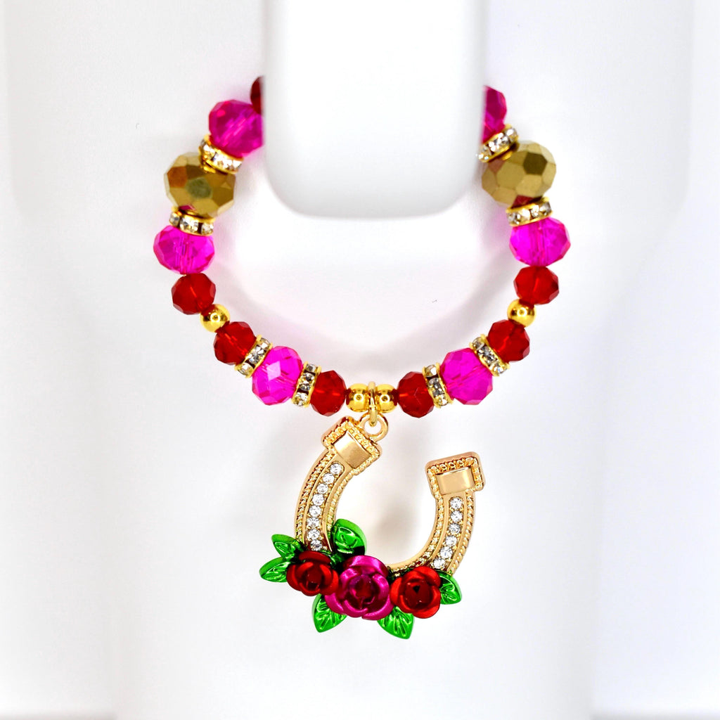 A tumbler handle beaded bracelet with a gold horseshoe charm. The tumbler bracelet features pink, red, and gold beads. The horseshoe charm is adorned with clear crystals and red roses with green leaves.