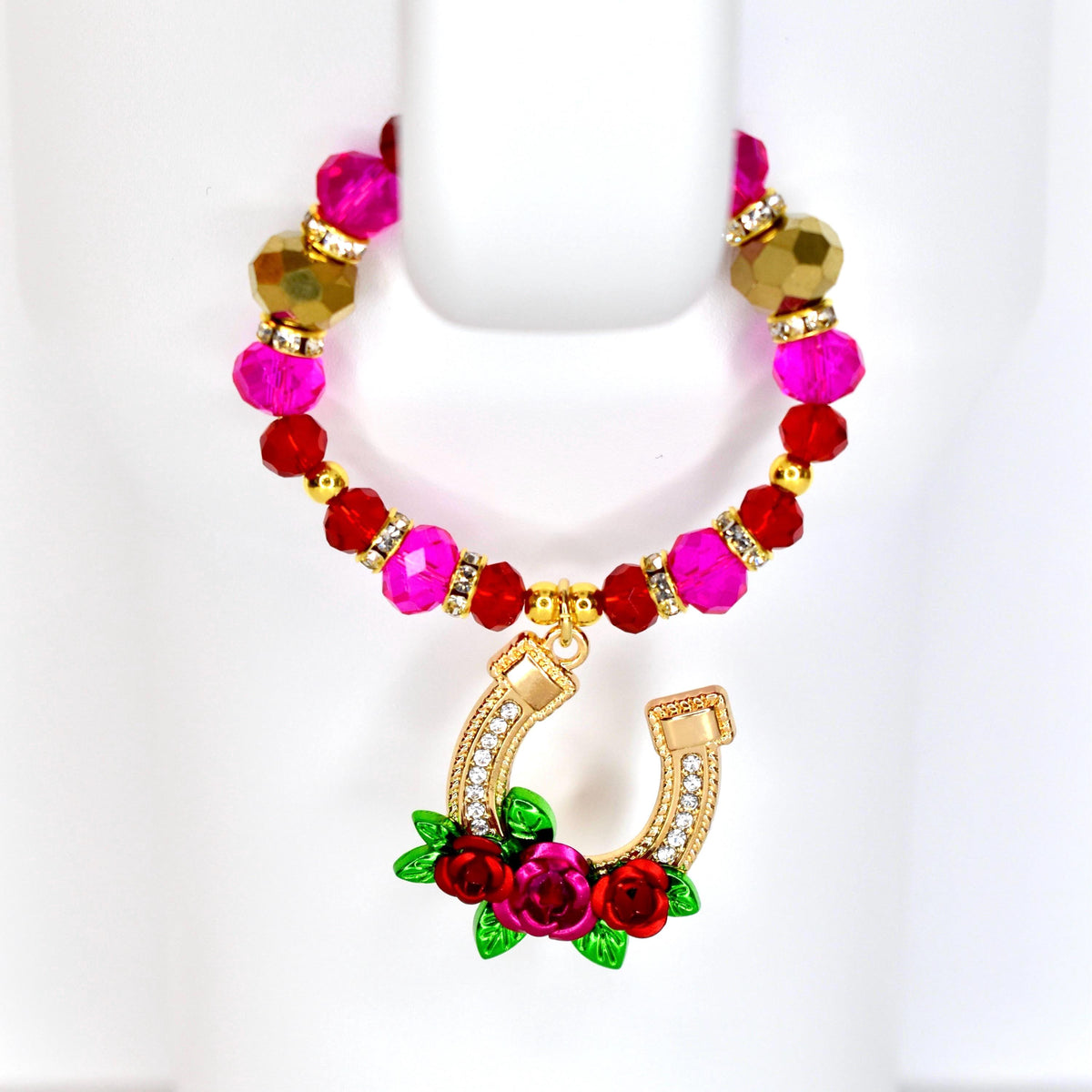 A tumbler handle beaded bracelet with a gold horseshoe charm. The tumbler bracelet features pink, red, and gold beads. The horseshoe charm is adorned with clear crystals and red roses with green leaves.