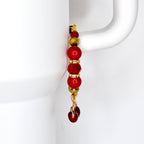 A tumbler handle charm with a gold-colored hook and chain. The charm features a series of red and gold beads, including round and faceted shapes. The bottom of the charm has a cherry charm.