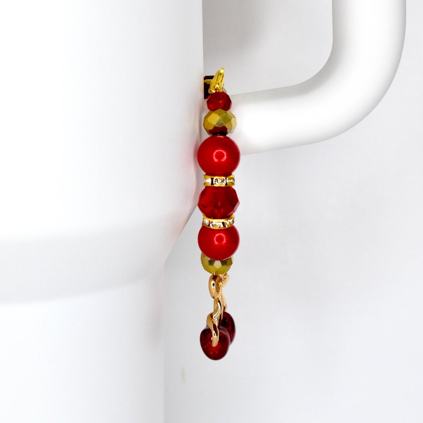 A tumbler handle charm with a gold-colored hook and chain. The charm features a series of red and gold beads, including round and faceted shapes. The bottom of the charm has a cherry charm.