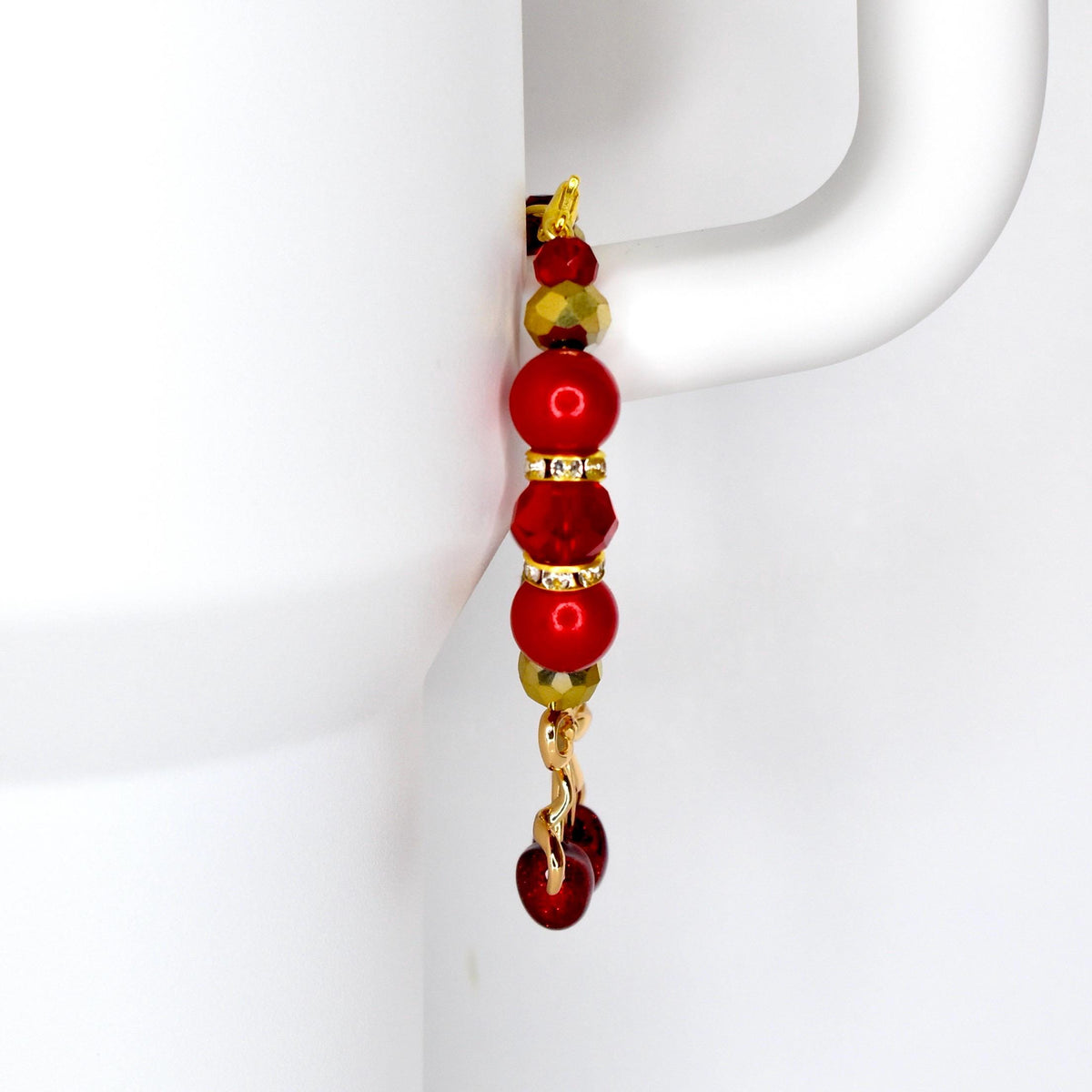 A tumbler handle charm with a gold-colored hook and chain. The charm features a series of red and gold beads, including round and faceted shapes. The bottom of the charm has a cherry charm.