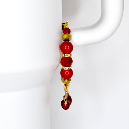 A tumbler handle charm with a gold-colored hook and chain. The charm features a series of red and gold beads, including round and faceted shapes. The bottom of the charm has a cherry charm.