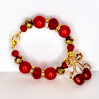 A red beaded bracelet with gold accents and a gold cherry charm. The bracelet features round red beads, faceted red beads, and gold-colored faceted beads. The charm is a gold bow with two red cherries.
