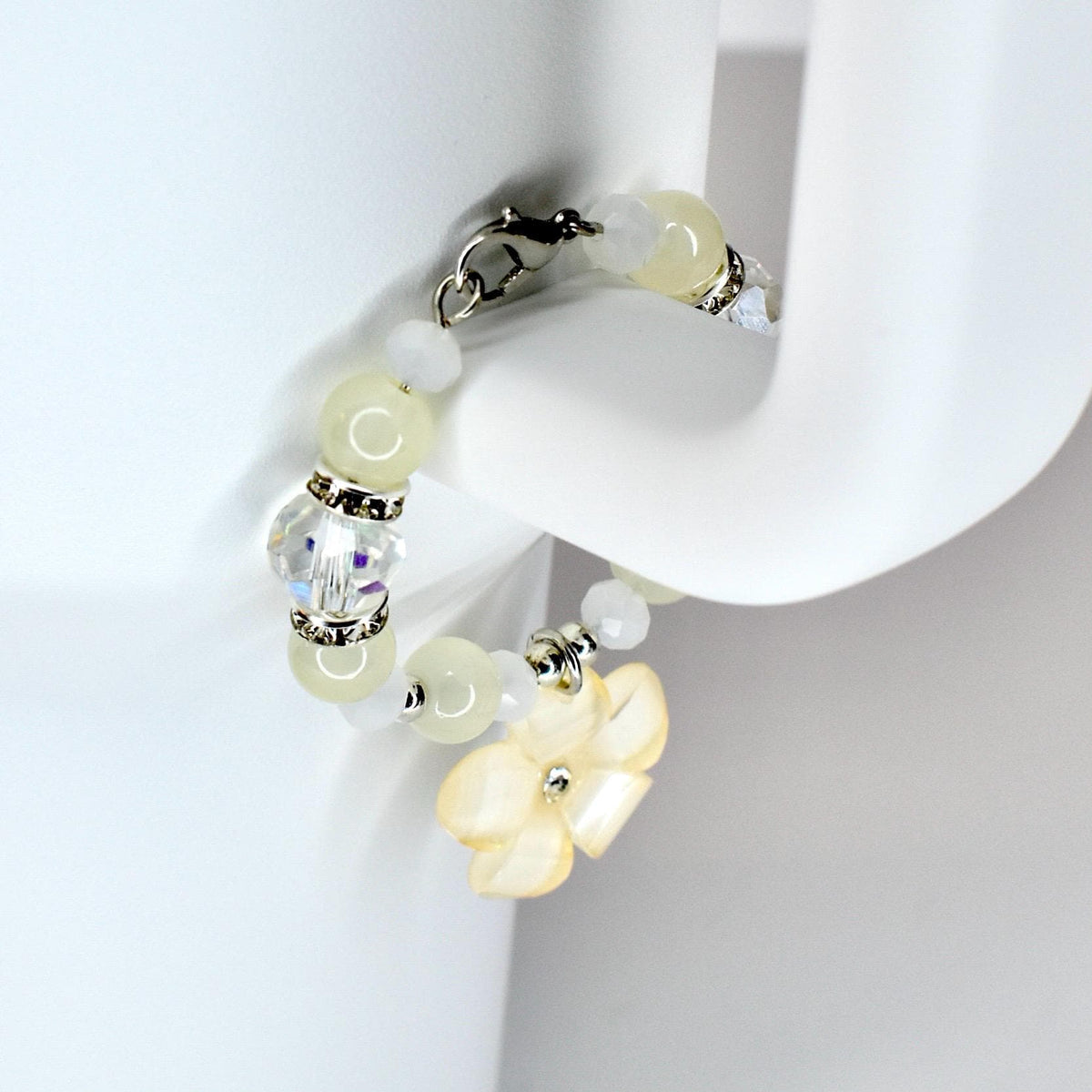 A delicate tumbler handle bracelet featuring a mix of pale yellow and white beads, accented with silver-toned metal. The bracelet includes a clear, faceted bead and a flower charm. The clasp is silver-toned.