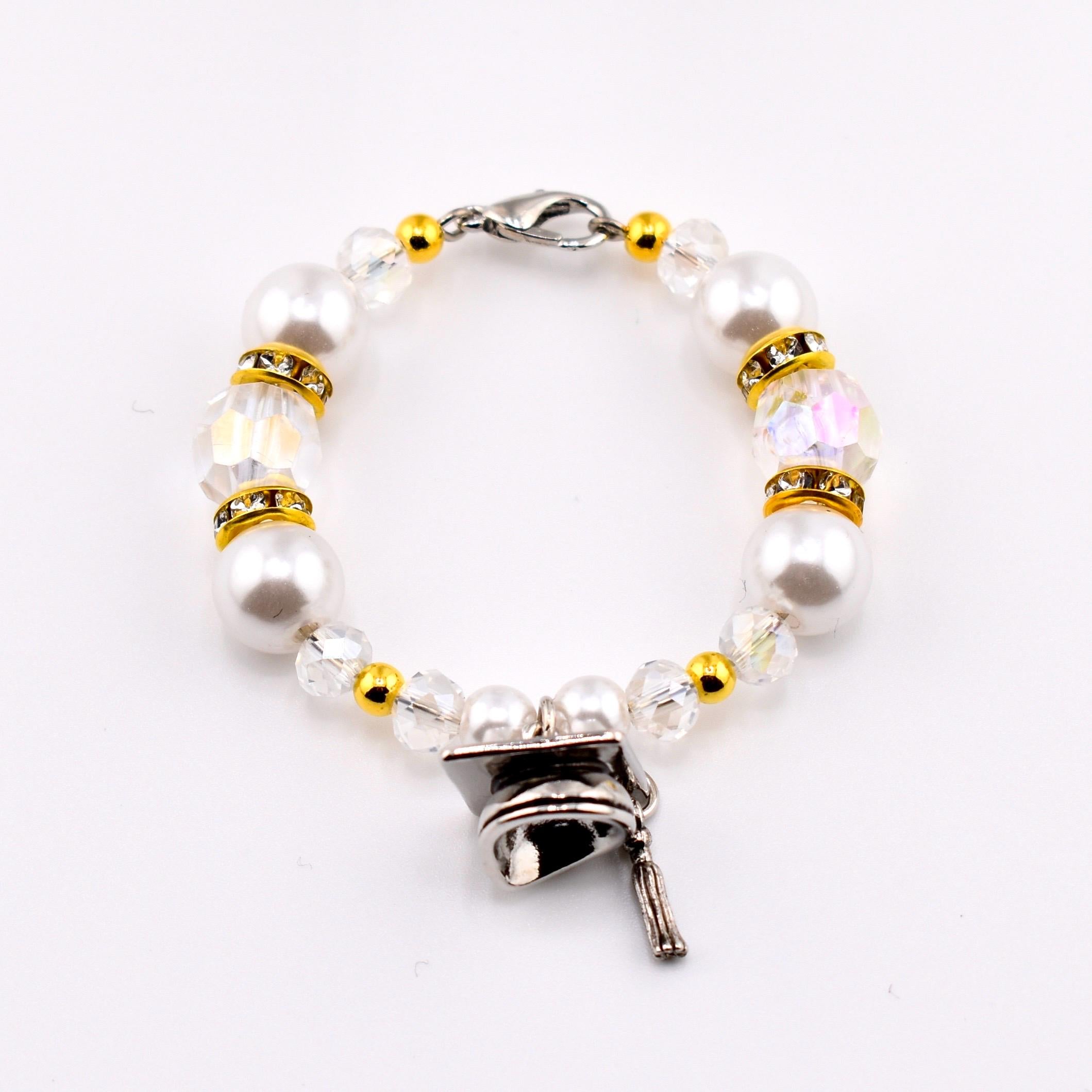 A beaded tumbler handle charm featuring large white faux pearls, clear iridescent beads, and gold-colored accents. The bracelet includes a silver-colored graduation cap charm with a tassel, perfect for a graduation gift.