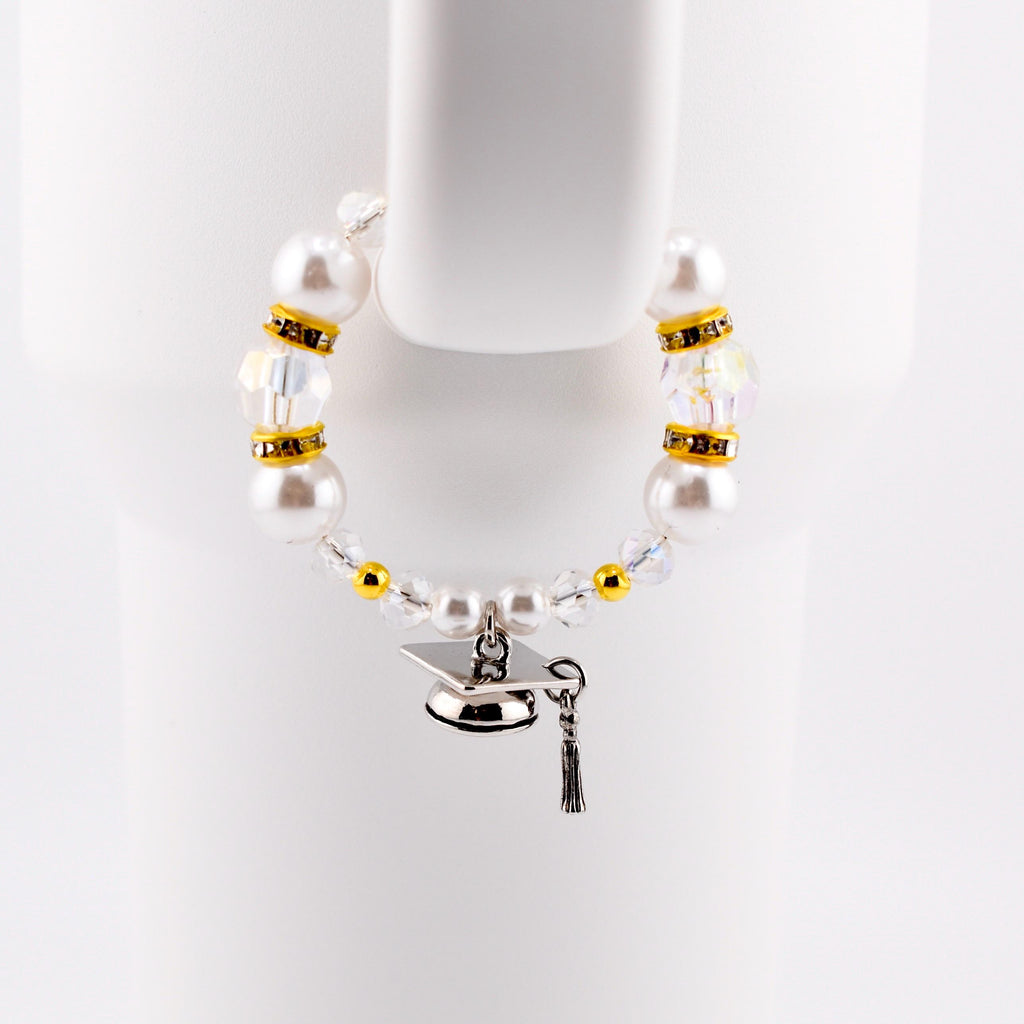 A graduation-themed tumbler handle bracelet featuring large white faux pearls, clear crystal beads, and gold accents. A silver graduation cap charm with a black tassel hangs from the bracelet. The bracelet is displayed against a white background.
