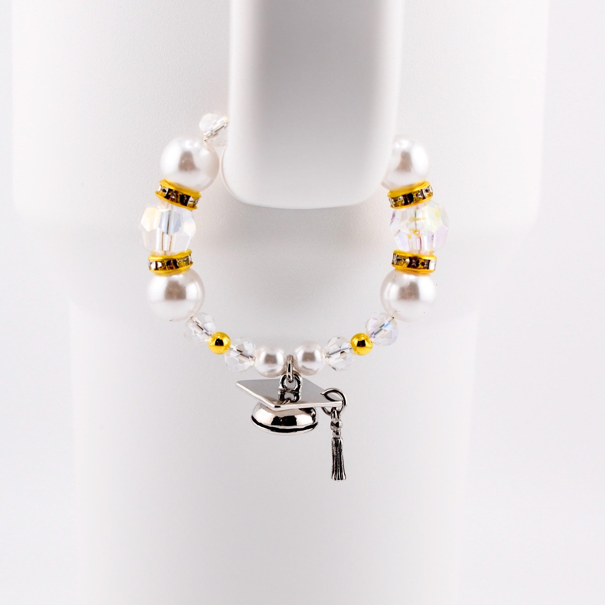A graduation-themed tumbler handle bracelet featuring large white faux pearls, clear crystal beads, and gold accents. A silver graduation cap charm with a black tassel hangs from the bracelet. The bracelet is displayed against a white background.