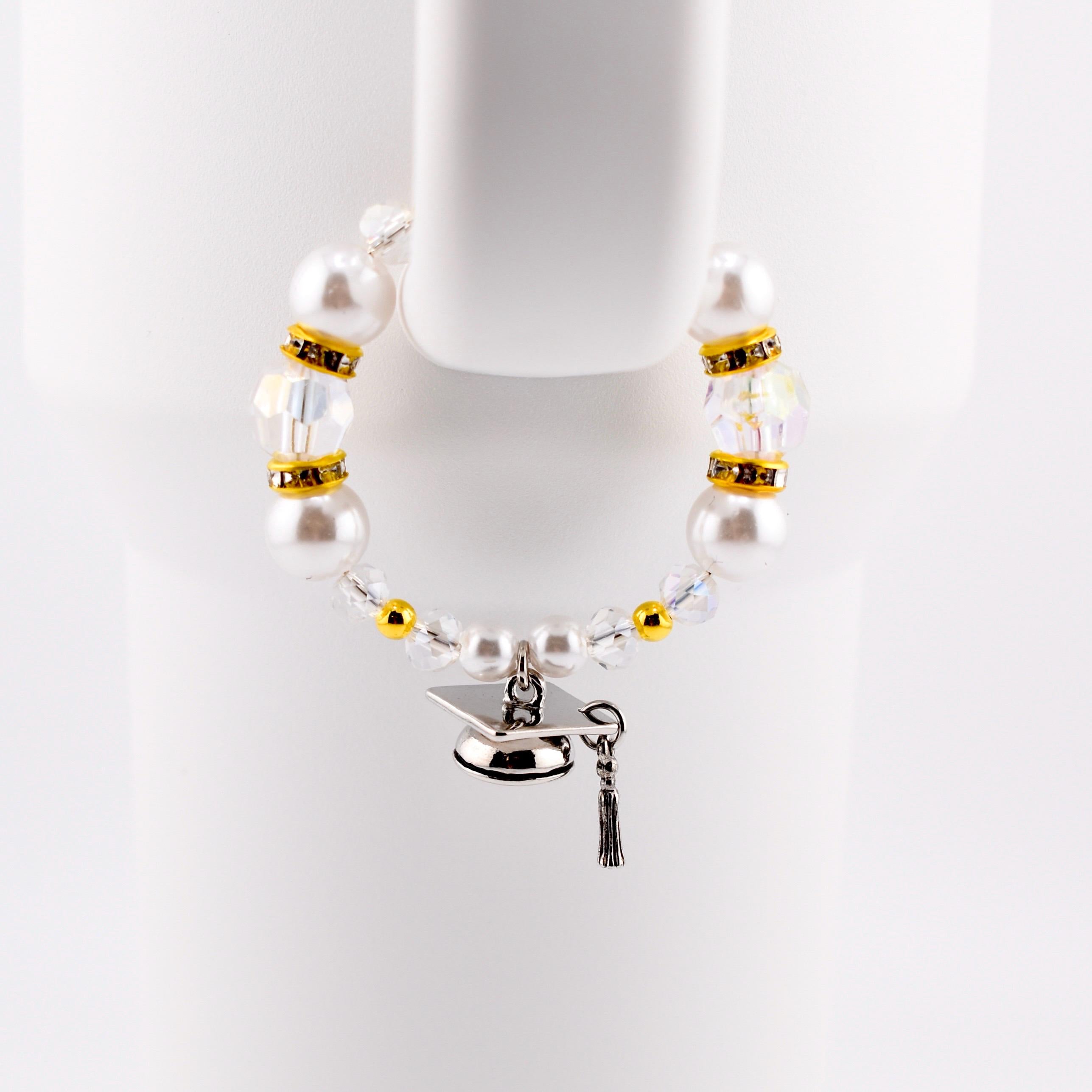 A graduation-themed tumbler handle bracelet featuring large white faux pearls, clear crystal beads, and gold accents. A silver graduation cap charm with a black tassel hangs from the bracelet. The bracelet is displayed against a white background.