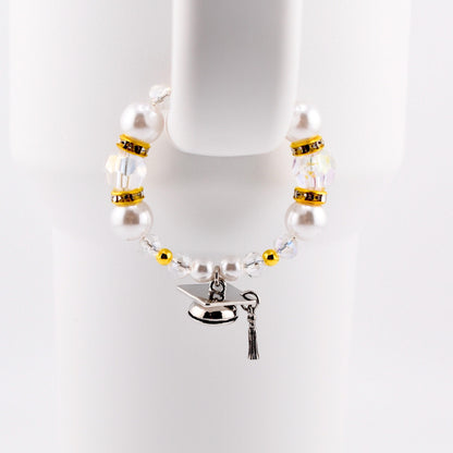 A graduation-themed tumbler handle bracelet featuring large white faux pearls, clear crystal beads, and gold accents. A silver graduation cap charm with a black tassel hangs from the bracelet. The bracelet is displayed against a white background.
