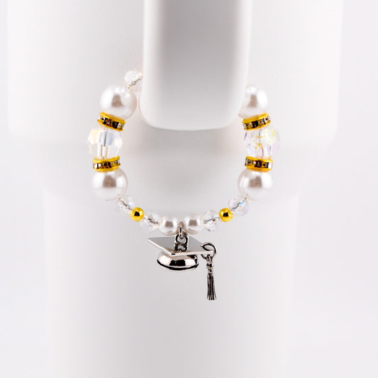 A graduation-themed tumbler handle bracelet featuring large white faux pearls, clear crystal beads, and gold accents. A silver graduation cap charm with a black tassel hangs from the bracelet. The bracelet is displayed against a white background.