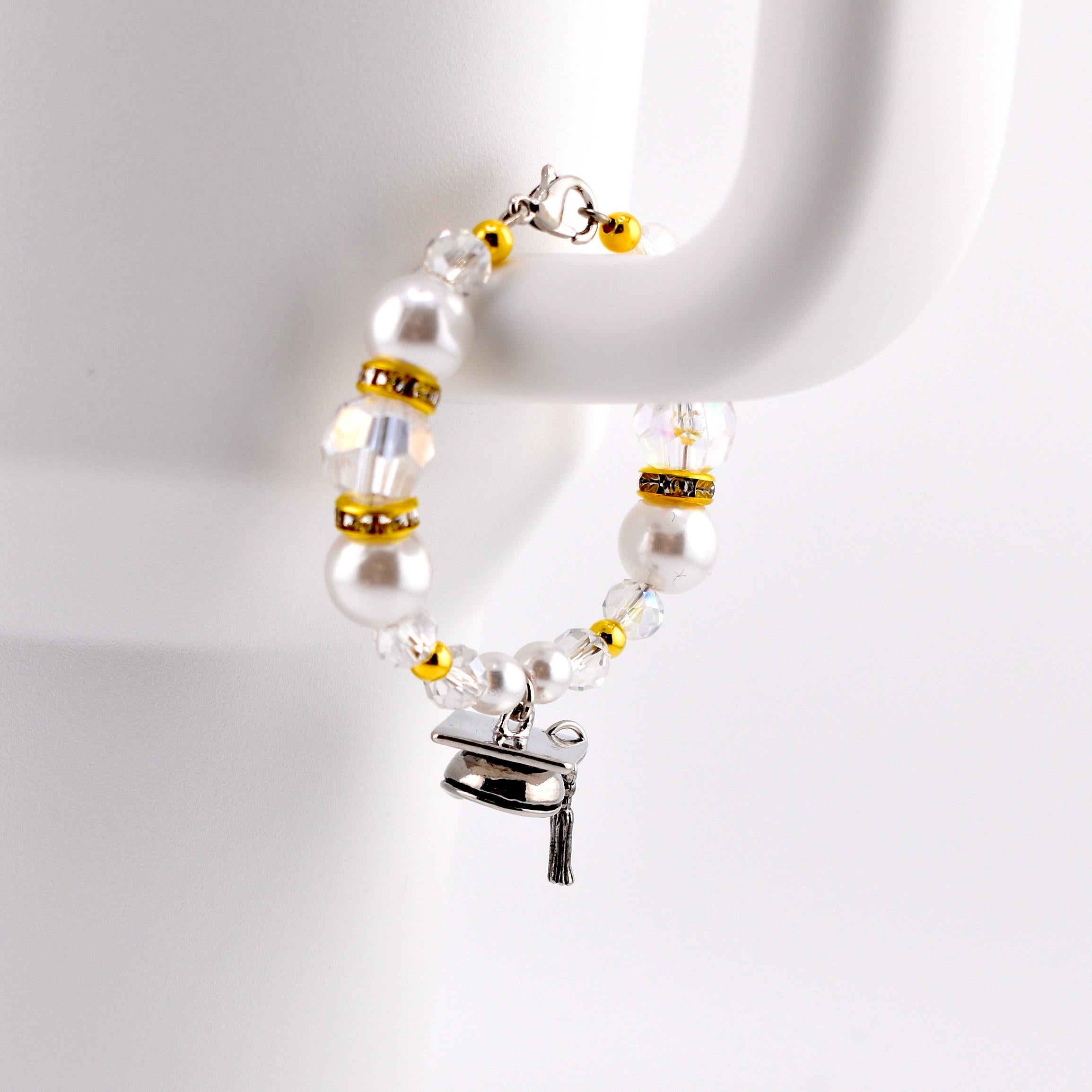 A white beaded handle charm with gold accents and a graduation cap charm. The tumbler handle bracelet features pearl-like beads, clear crystal beads, and gold-colored spacers. The graduation cap charm is silver with a black tassel.